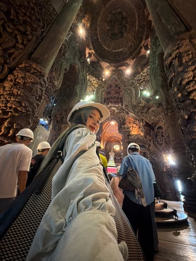 The image depicts the interior of the Sanctuary of Truth in Pattaya City, Thailand, a monumental wooden structure intricately carved with religious and mythological motifs. In the foreground, a young woman wearing a light-colored headscarf and a white hat is captured in a selfie-style pose, looking over her shoulder at the camera. She is dressed in a loose, light-colored top. To her left, two individuals are visible wearing white hard hats, suggesting they might be workers or visitors. One of them is wearing a white t-shirt and grey pants, while the other is wearing a black t-shirt. Further into the background, a person in a blue shirt and black pants is seen walking away from the camera. The environment is characterized by massive, ornately carved wooden columns and ceilings, illuminated by spotlights that highlight the detailed artwork. The overall atmosphere suggests a tourist or cultural site, with the architecture being the primary focus of the scene, interspersed with the presence of people.
