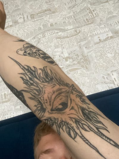 This media file is a close-up, eye-level shot of a person's arm, which is adorned with several black ink tattoos. The main tattoo prominently features an abstract, eye-like motif with sharp, spiky elements radiating outwards, reminiscent of a stylized sun or creature's headdress. To the left of this central design, another tattoo is partially visible, appearing to depict a skeletal figure or a mythological creature with intricate details. The person's skin is fair, and there is visible hair growth on the arm.
The background consists of patterned wallpaper, featuring a repetitive design of architectural elements, possibly buildings or cityscapes, in light grey and white tones. A dark blue fabric, likely clothing, is visible in the upper right portion of the frame, partially obscuring the background and the person's body. The lighting appears to be artificial or from an overcast day, casting a soft, diffused light on the scene. The focus is sharp on the tattoos, while the background is slightly softer.
The image seems to be taken indoors, in a private setting, possibly a home. There is no discernible time of day or weather information. The emotions conveyed are neutral, with the focus solely on the visual details of the tattoos and their immediate surroundings. No text is visible in the image. The location context provided is Severodvinsk, Russia, but this cannot be independently verified from the visual content alone.