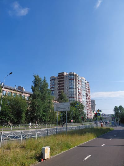 The media file shows an outdoor scene on a bright, sunny day with a clear blue sky. A tall, modern residential building with red and white accents dominates the left side of the frame. In front of this building, a sidewalk is lined with a metal fence and green grass. A paved road runs alongside the sidewalk, with white markings indicating lanes.
Several traffic signs are visible. One sign, with an arrow pointing left, reads "проспект Непокоренных 0,3" and below that "улица Хлопина 0.8". Another sign indicates a pedestrian crossing. Traffic lights are also present, with one showing red.
On the sidewalk, two people can be seen walking. One appears to be wearing shorts and a t-shirt, while the other is wearing a white shirt and shorts. Further down the road, a few cars are parked or driving. A metal box, likely an electrical or utility enclosure, sits near the side of the road on the grassy area. The scene suggests a typical urban environment in what appears to be a residential area.