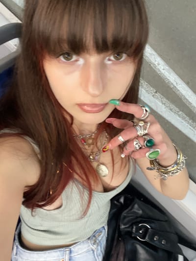 A female subject is depicted in a selfie-style portrait, looking directly at the camera. She has long, straight, reddish-brown hair with bangs, brown eyes, and light pink lips. White eyeliner is applied to the inner corners of her eyes. She is wearing a light green ribbed tank top and light blue denim jeans. Several layered silver-toned necklaces are visible, one featuring a large coin pendant and another with a Star of David charm. Small gold-toned glitter strands are present in her hair. Her left hand is raised, with the index finger lightly touching her chin or lower lip. This hand is extensively adorned with numerous silver-toned rings, including several with green stones (one large oval, one small round), and multiple silver and gold-toned bracelets on her wrist. Her fingernails are long, with green polish on the index finger and other varying designs. A black leather-like bag with a silver buckle is positioned on her right side. The background to the right displays a light-colored surface with distinct raised white horizontal lines, while the upper left shows dark, possibly upholstered seating. There are no specific identifying markers within the image to confirm a precise location within the city of Raanana, Israel.