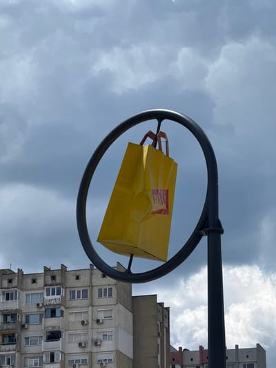 A yellow shopping bag sculpture hangs within a dark circular frame, mounted on a black pole, against a cloudy sky. The sculpture features a red logo with stylized letters. In the background, a multi-story, utilitarian apartment building with many windows and air conditioning units is visible, a common sight in Shumen, Bulgaria. The building's facade shows signs of wear. Further in the background, more buildings with varying architectural styles can be seen. The overall impression is one of an urban environment under overcast weather.