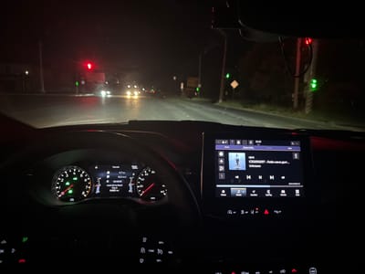 The image shows the interior of a car at night, with the driver's perspective looking forward. The car is on a wet road, indicating recent rain. Traffic lights are visible ahead, with a red light on one and green lights on another. The car's dashboard is illuminated, showing the speedometer, tachometer, and a digital display. The central infotainment screen is also on, displaying music playback information. The scene suggests the car is stopped at a traffic light or moving slowly in urban traffic. The presence of a "Poltava" license plate on the car confirms the location.