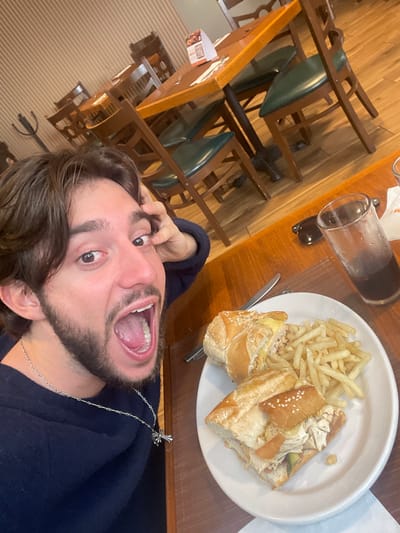 A young man with dark, medium-length hair and a beard is taking a selfie in a restaurant. He is wearing a dark blue sweater and a silver cross necklace. His mouth is open in a surprised expression, and he is touching his head with his right hand. In front of him on a wooden table is a white plate with a sandwich cut in half and a pile of french fries. To the right of the plate is a glass of dark liquid, likely soda, and a pair of dark sunglasses. The background shows a dining area with wooden tables and chairs, and a wall with horizontal wood paneling. The restaurant appears to be located in Cuautitlán Izcalli, Mexico.
