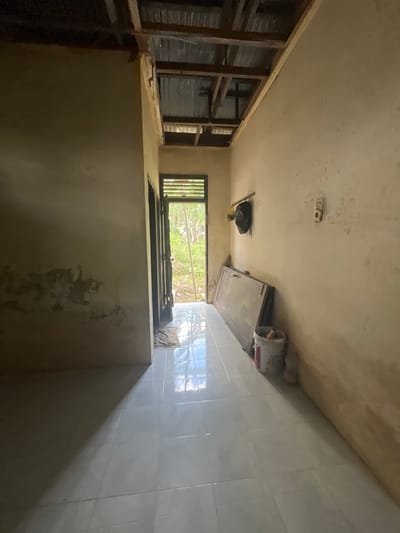 The image depicts an interior view of a room under renovation in Kota Kuala Simpang, Indonesia. The room is a narrow hallway with a white tiled floor. The walls are painted a light yellowish-beige and show signs of wear, with patches of discoloration and what appears to be water damage. The ceiling is incomplete, with exposed wooden beams and corrugated metal sheeting visible.

In the background, an open doorway leads to an outdoor area with lush green vegetation. A dark-colored object, possibly a bag, hangs on the wall to the left of the doorway. To the right of the doorway, a large, flat object, possibly a door or a piece of furniture, leans against the wall. A white bucket with red accents, containing tools or materials, sits on the floor next to it. There is a light switch or outlet on the wall to the right.

The lighting suggests it is daytime, with natural light streaming in from the doorway and reflecting off the tiled floor, creating strong shadows. The overall impression is one of an unfinished or in-progress construction or repair project. There are no people visible in the image.