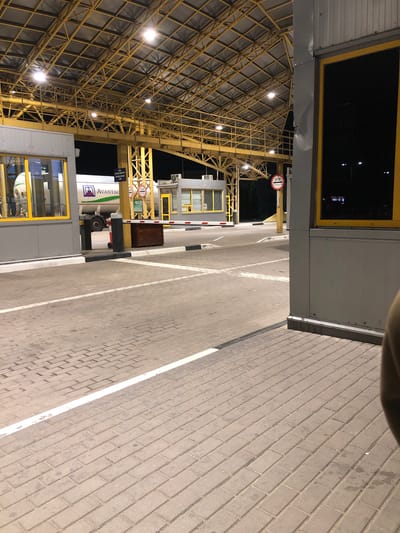 The image shows a border crossing checkpoint at night, likely at the Starovoitove, Ukraine location given the context. The scene is brightly lit by overhead fluorescent lights under a large, yellow metal canopy. In the foreground, there is a paved area made of interlocking bricks with white lines marking lanes. A raised white curb runs along the edge of the paved area.
To the left, there is a grey booth with yellow-framed windows. A truck with a white tanker trailer bearing the "AVANTAGR" logo is visible through the window of another booth further back. A red and white striped barrier arm is lowered across a lane.
On the right side of the image, a grey structure with a yellow-framed window is partially visible in the foreground, with a person's shoulder and arm appearing on the far right edge of the frame. A red circular sign with "STOP" and Cyrillic text is mounted on a yellow pole. Several other smaller buildings and structures are visible in the background, including what appears to be a control booth. The overall environment suggests a functional, active border crossing.