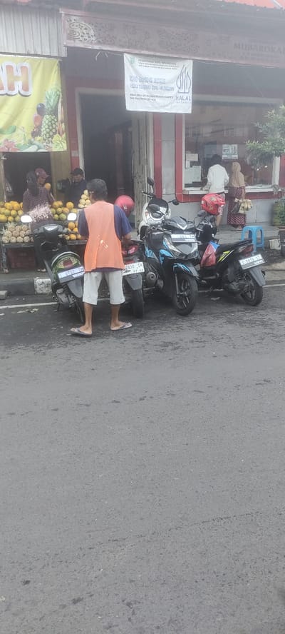 The scene is a street in Mojokerto, Indonesia, located at latitude -7.467 and longitude 112.438. There are three motorcycles parked on the street, one of which is a blue and black Honda Vario with the license plate S 6881 TM. A man wearing an orange vest and white pants is standing next to the motorcycles, facing the street. Behind the motorcycles, on the sidewalk, there is a small store selling fruit and vegetables. There are at least three people inside the store, two of whom are women, one of whom is wearing a blue shirt and blue pants, and another wearing a black shirt and white pants. The shop window has a sign that says "KIOS DAGING MURAH PASAR TINGGIK 2018 MOJOKERTO". There is also a sign on the shop that says "HALAL". There is another sign on the wall above the shop that says "MUBAROKAH". The street is made of asphalt and is empty of traffic. The sky is overcast and grey.