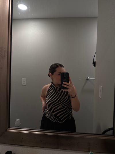 A person stands in front of a bathroom mirror holding a phone. The mirror frame is gold-toned. The person wears a zebra-striped top and a black skirt. The phone has visible initials on the case, and the person also wears bracelets. The bathroom has a light, a silver-toned faucet, and a wall outlet.