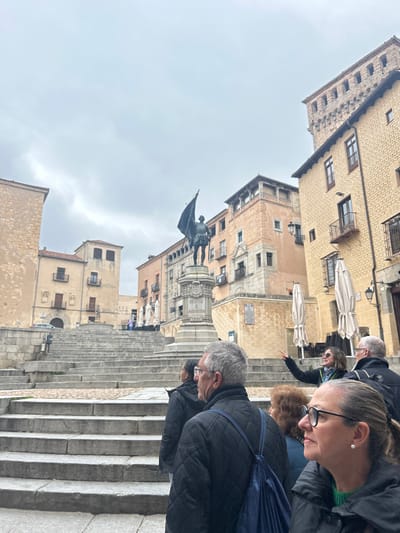 The image shows a group of tourists standing on a set of stairs in the Plaza Mayor in the historic city of Segovia, Spain. The plaza is located at coordinates 40.949, -4.122. The group is looking at a statue of a man holding a flag, which is located in the center of the plaza on a stone pedestal. The statue is surrounded by a large cobblestone plaza with multiple sets of stone stairs. The plaza is bordered by old buildings, some of which have intricate facades and balconies. The sky is overcast and gray. There are two women standing in the foreground, and a man with a blue backpack is standing in front of them. A woman in a white shirt is standing at the bottom of the stairs pointing toward the statue. Another man is standing behind her. The woman in front is wearing a green and black sweater and glasses. The woman behind her is wearing a black jacket and has a hand on her hip. The man with the backpack is wearing a black jacket and a blue backpack. The man standing behind her is wearing a black jacket and is looking at the statue.