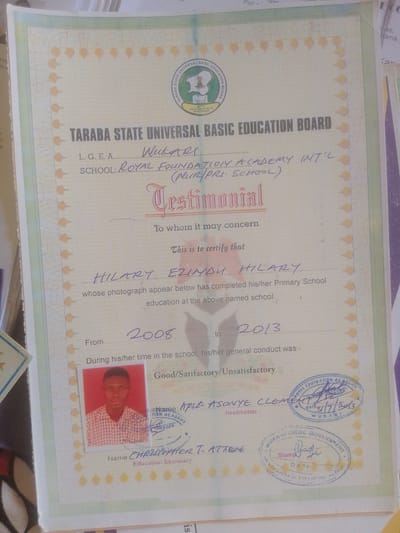 This is a photograph of a testimonial document from the Taraba State Universal Basic Education Board. The document, dated 2013, is for a student named Hilary Ezimadu Hilary, who completed their primary school education at Royal Foundation Academy Int'l (Nuryrri School) in Wukari. The document includes a photograph of the student, a signature of the Headmaster, Apsu Asonye Clement, and stamps from the Royal Foundation Academy and Wukari Local Government. The specific location within the city of Magajin Gari, Nigeria, is not mentioned on the document.