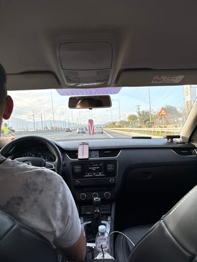 The image depicts the interior of a vehicle, viewed from a rear passenger seat looking forward through the windshield onto a highway in Koropi, Greece.
**Vehicle Interior:**
The driver, visible from the back of the head and right ear, is a male wearing a light-colored, possibly patterned, short-sleeved shirt. His hands are positioned on a black Skoda steering wheel, where the dashboard display shows a speed of "65," likely indicating 65 km/h. A smartphone (iPhone) is mounted centrally on the dashboard, displaying what appears to be a navigation or mapping application. Below it, the car's infotainment system is active, displaying "Υποκλοπη-Bluetooth" and "iPhone," indicating a Bluetooth connection. Below the infotainment system are controls for climate and other functions. A manual gear shifter is present in the center console. A plastic bottle of "Vikos" brand water is situated in a cup holder in the center console, connected to a white charging cable. A small electronic device with red indicator lights is visible near the gear shifter. The front passenger seat, upholstered in black leather, is unoccupied. The rearview mirror reflects a portion of the driver's head and has a pink and white "V"-shaped air freshener hanging from it. The passenger-side sun visor is raised and displays an "AIRBAG" warning label. The vehicle's headliner is light grey.
**Exterior Environment:**
Through the windshield, a multi-lane highway is visible, with several other vehicles traveling in the same direction. The highway features streetlights and utility poles lining its sides. On the right-hand side of the road, there is a paved shoulder, followed by a grassy verge and trees. A yellow, triangular warning sign depicting road workers is clearly visible on the right side of the highway, along with a circular regulatory sign. In the distant background, an elevated structure, possibly a railway or an overpass, with visible supporting pillars, spans horizontally across the landscape. The sky is clear blue with minimal cloud cover, indicating daytime conditions.