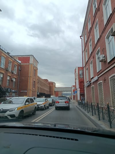 The image captures a street scene in Orekhovo-Zuyevo, Russia, on an overcast day. The perspective is from inside a vehicle, looking forward through the windshield. The road is filled with a line of cars, suggesting a traffic jam or a slow-moving procession.
Dominating the right side of the frame is a large, red brick building with multiple floors and numerous windows. White trim frames the windows, and several air conditioning units are visible mounted on the exterior. The building's architecture appears industrial or utilitarian. On the left side of the street, another similar red brick building stretches into the distance, featuring more windows and visible architectural details like pipes and external conduits. Some sections of this building show signs of wear, with faded red paint.
Several vehicles are identifiable in the traffic. In the foreground, a white sedan with yellow and black markings, likely a taxi, is prominent. Other cars, including a silver SUV with a visible license plate, are part of the queue. The license plate reads "P036KH 750".
Visible text includes "КУХНИ" (KITCHENS) on one of the buildings on the left, indicating a business. Another sign on the left reads "АВЕ" and "СМОУК". A blue parking sign is also visible on the right side of the street.
A pedestrian, dressed in dark clothing and carrying a bag, is walking along the sidewalk on the right, past a decorative black metal fence. A pedestrian bridge spans the street in the distance, connecting the buildings. The overall mood is neutral, reflecting the mundane activity of city traffic under a cloudy sky. The time of day is difficult to pinpoint precisely due to the overcast conditions but appears to be daytime.