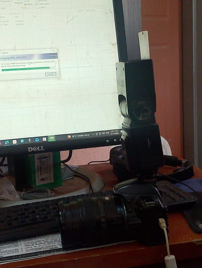 The image captures a close-up view of a desktop computer setup in what appears to be a residential or office environment in Kubwa, Nigeria. A Dell monitor displays a "Copying DSC_0293.JPG" progress window, indicating a file transfer is in progress. Below the monitor, a black keyboard is visible, and to its right sits a digital camera, likely a DSLR, with a lens attached. A flash unit is mounted on the camera's hot shoe, and a white cable is plugged into the camera. The background features a reddish-brown wall and part of a door. The scene suggests a digital photography workflow, possibly related to personal or professional use, taking place in an indoor setting in Kubwa.
