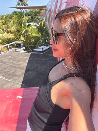 This is a close-up, side-profile photograph of a young woman with long, dark auburn hair illuminated by sunlight. She is wearing sunglasses with tortoise-shell frames and a silver chain necklace. Her head is tilted, and she appears to be resting on something pink and textured, possibly a lounge chair or the side of a structure.

The background suggests a tropical setting with lush green palm trees and foliage visible to her left. A gray, weathered surface, possibly pavement or a roof, occupies the right side of the image, with a stark shadow cast across it, indicating a sunny day. A metal ladder or staircase is also visible in the background, leading up towards the foliage and what appears to be a building.

The overall mood is relaxed and casual, with the bright sunlight and tropical greenery suggesting a warm, outdoor environment. The focus is on the woman and her immediate surroundings, with the backdrop providing context of a potentially resort-like or coastal location in Blahbatuh, Indonesia. The image is taken from a low angle, looking up at the woman. There is no visible text in the image.
