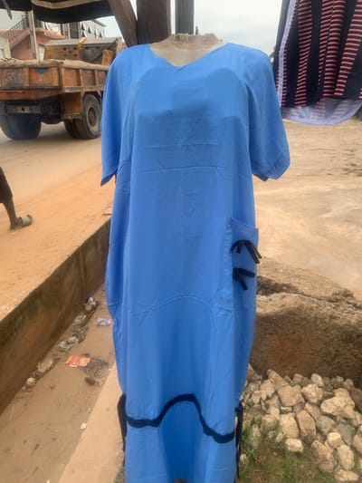 A blue, v-neck, ankle-length dress is displayed on a mannequin outside a store in Keffi, Nigeria. The dress has short sleeves, two black-ribbon-adorned pockets on the right side, and a black trim at the hem. Behind the dress, a yellow dump truck is parked on a dirt road. To the left, a person wearing a black t-shirt and green flip-flops walks away from the camera. The ground is sandy and rocky, with some scattered debris. The overall scene suggests an outdoor market or roadside stall.