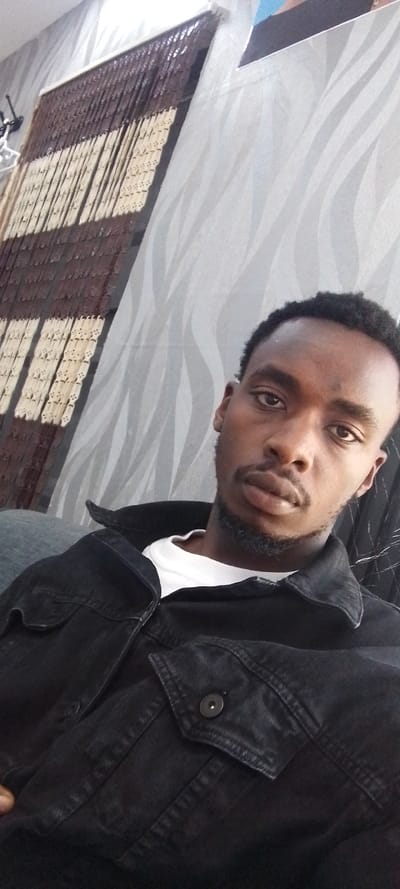 A male individual is depicted in a close-up, head-and-torso portrait within an indoor setting in Matathani ward, Kenya. The subject has short, dark, curly hair, a short beard, and a mustache, and is looking directly at the camera with a neutral expression. He wears a white long-sleeved crew-neck shirt beneath an open black denim jacket. The jacket features a collar, two visible chest pockets with buttoned flaps, and dark buttons. The background to the subject's left consists of a multi-toned beaded curtain, primarily dark brown and light tan, hanging from a white rail. To the subject's right, a wall is covered with light gray wallpaper displaying a repeating wavy pattern on a lighter gray base. The image is taken at a slight upward angle, with indoor lighting illuminating the scene.