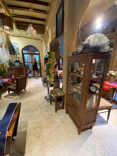 The image depicts the interior of a dimly lit cafe with a rustic and vintage ambiance, situated in Mũi Né, Vietnam. The walls are a textured yellow, with exposed wooden beams on the ceiling. Various vintage items are displayed throughout the space. In the foreground, a wooden bench with a blue cushion is partially visible on the left. To the right, a tall, glass-fronted wooden cabinet displays an assortment of antique objects, including clocks, teacups, and what appear to be small ornaments. A standing fan is positioned in front of the cabinet. Further to the right, a wooden stool with a tan seat is partially visible. In the mid-ground, a woman in a black dress stands near a wooden doorway, holding a phone. Another woman is visible in the background to the right, near a table. The floor is a speckled concrete. The overall impression is that of a quaint, retro establishment with a collection of decorative vintage pieces.