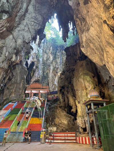 The image captures the majestic interior of a large natural cave, featuring a vibrant man-made staircase and religious structures. Natural daylight streams in from a vast opening in the cave ceiling, illuminating the scene.

In the foreground and midground, a wide, curving staircase dominates the view. Its steps are painted in bold, horizontal stripes of red, orange, yellow, green, and blue, creating a striking visual contrast with the natural rock formations. Several people are seen on these stairs, either walking up or down, standing, or observing their surroundings, suggesting they are visitors or pilgrims exploring the site.

The cave walls are characterized by rugged, textured rock, displaying various shades of grey, brown, and off-white. On a rock ledge to the right, a brightly colored Hindu shrine or altar is visible, intricately decorated and featuring a multi-tiered dome. Further into the cave, above the stairs, another small building with a blue sign is nestled against the cave wall. Text in an Indian script, along with the partially visible English words "SRI VALLI DEVAN" and "15th," can be seen on the signs. Metal railings run alongside the staircase, and a green grated structure is located in the upper right portion of the image. The scene conveys a sense of awe and activity within a significant natural and religious landmark.