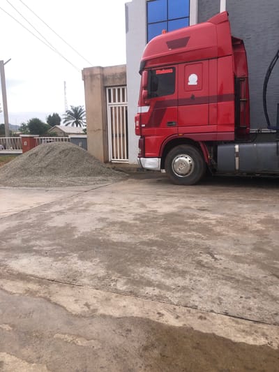 In New Stadium, Nigeria, a bright red truck is parked beside a building with a white and tan facade. The truck's cab features the name "Maduwa" in red lettering and a circular emblem. A large pile of gravel sits in front of the building, near a white gate. The ground is a concrete surface that appears wet and stained in patches. In the background, trees and a cell tower are visible against a cloudy sky. The overall scene suggests an industrial or construction-related location.