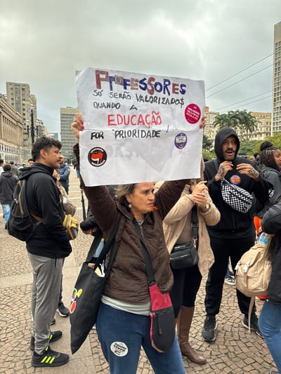 A medium shot shows a group of protestors holding a sign in Portuguese reading "PROFESSORES, SÓ SERÃO VALORIZADO$ QUANDO A EDUCAÇÃO FOR PRIORIDADE!" (TEACHERS WILL ONLY BE VALUED WHEN EDUCATION IS A PRIORITY!). The sign also includes stickers and political slogans.

The protestors are diverse in age and dress. Some are wearing casual clothing, while others have political stickers on their clothing and bags. The background features buildings and street elements, likely in a public square or street setting. The scene appears to be a political demonstration or protest. Based on the buildings and cobblestone ground, the location is likely in the city of São Paulo, Brazil.