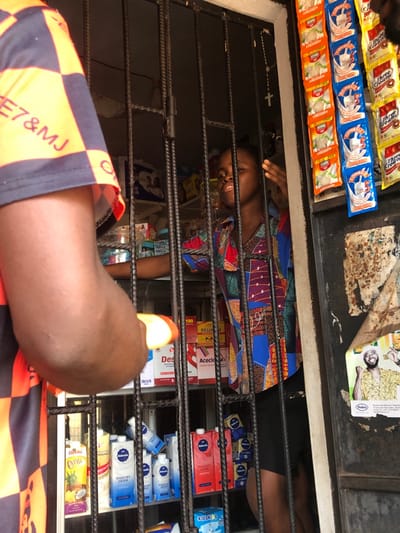 Through a metal-barred window, a woman in a colorful patterned shirt is visible behind shelves stocked with various items. These include beverage cartons like "Chivita" and "Kisski", dairy products labeled "Yogurt Drink", and boxes of "Acecloa" and "Rellx." On the outside of the window are products like "Three Crowns" milk powder. A leg in checkered orange and black shorts is positioned in the foreground, partially obscuring the view.