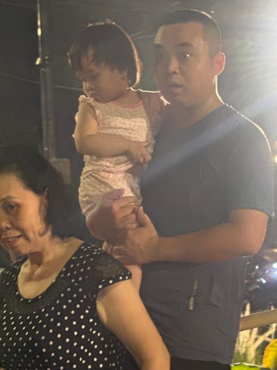 An outdoor, nighttime image shows a Vietnamese family at an event. A man in a dark t-shirt holds a young child wearing a pink outfit. To the left, a woman with dark hair wears a black top with white polka dots. Bright lights are visible in the background, suggesting the event is illuminated. The scene is likely in Hanoi, Vietnam, given the presence of what appears to be a Vietnamese family.