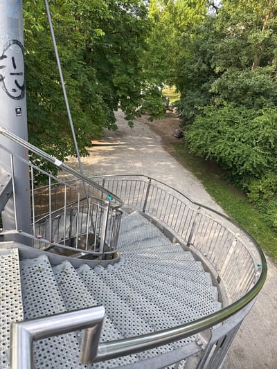 This is a high-angle, eye-level shot of a spiral staircase made of metal. The staircase has a curved railing made of brushed metal, and the steps are perforated with circular holes. The stairs are empty, and no activity is taking place.

The staircase is set in an outdoor park-like environment. To the left of the stairs, lush green trees and bushes are visible, casting dappled shadows on a dirt path. In the distance, the path leads towards a wooded area with more trees. A few scattered leaves are on the steps of the staircase, suggesting it's likely autumn or a time when leaves fall. The lighting suggests it's daytime, with sunlight filtering through the trees. The scene appears calm and peaceful.

There are some graffiti tags on the large cylindrical pole supporting the staircase in the lower left corner of the image. One tag appears to be a black smiley face with a tongue sticking out, and another sticker with the text "CTAZIDE" and "PMSS" is visible.

The overall scene depicts a public outdoor space, possibly a park or recreational area, featuring a modern metal spiral staircase integrated into a natural landscape.