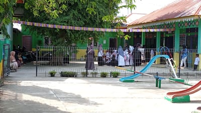In Uteunreutoh, Indonesia, a group of children in traditional Indonesian attire are gathered on a school grounds, seemingly waiting for an activity to begin. Some children are accompanied by adults, while others appear to be supervised by teachers. The school building is visible in the background, with its distinctive green walls and orange roof. A playground with a slide and seesaw is also present, suggesting a space for recreation and play. The atmosphere appears calm and orderly, with a sense of community and shared purpose.