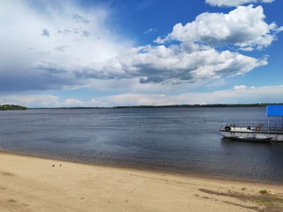 The image shows a wide, calm river with a sandy beach in the foreground. The river is dark blue and appears to be very deep. A small boat is docked at a pier on the right side of the image. The pier has a blue roof and a railing. The beach is light brown and appears to be made of sand and small stones. There are a few birds on the beach. In the background, there is a line of trees on the opposite bank of the river, and a blue sky with white clouds. The scene is peaceful and serene. The location is the Volga River embankment in Volsk, Russia.
