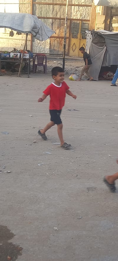 In Khan Yunis, Palestinian Territories, a young boy in a red t-shirt and dark shorts runs across a dusty, uneven ground. He wears dark sandals. In the background, several makeshift structures are visible, including a tent-like shelter with a grey tarp and a market stall covered by a white tarp, displaying boxes of goods. A red plastic chair is positioned near the market stall. Other individuals, including a boy in a black t-shirt and tan pants, are present in the background, one partially obscured by the tent. The environment appears to be an outdoor area with debris scattered on the ground.