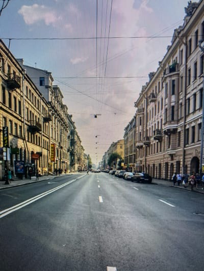 The image depicts a wide city street lined with tall, multi-story buildings on both sides. The buildings appear to be old, with classical architectural details, and are painted in light colors. There are balconies on some of the buildings. The street is paved and has double white lines marking lanes. Several cars are parked along the side of the road on the right. Pedestrians are visible walking on the sidewalks on both sides of the street. Overhead, there are numerous electrical wires crisscrossing the sky, attached to poles and buildings. The sky is partially cloudy with a light blue hue. The scene suggests a day in an urban environment. The provided context of Naberezhnye Chelny, Russia, is not evident from visual cues within the image itself, as there are no discernible landmarks, signs, or architectural styles that specifically identify this location.