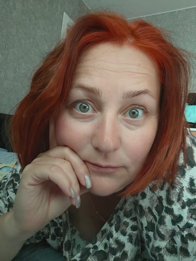 A close-up shot depicts a woman with red hair, gray eyes, and fair skin. She is wearing a leopard-print top. Her left hand is positioned against her face, with her fingers near her mouth. The background appears to be a wall and a glimpse of a bed. The image location in the city of Markovskii, Russia, is unidentifiable from visual evidence.