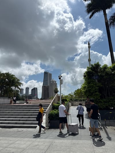 This image captures an outdoor scene with several people, lush greenery, and modern buildings under a cloudy sky. The perspective is somewhat skewed, making it appear as if the ground is vertical.
In the foreground, a person in a white t-shirt and black shorts is walking down a wide set of stairs, casting a distinct shadow. Further up the stairs, another person wearing a yellow and black striped shirt and dark pants is also walking. A few other individuals are visible on the stairs and the walkway. One person is pulling a light-colored rolling suitcase, and another is carrying a dark bag.
The setting appears to be a public space with well-maintained stairs and a paved walkway. Behind the people and stairs, dense green trees and foliage create a natural backdrop. In the distance, several tall buildings, characteristic of a city skyline, are visible against a dynamic sky filled with dramatic, dark clouds interspersed with patches of blue. A tall pole with a camera or sensor is also present in the frame. The overall impression is that of a busy urban environment on a day with changeable weather, possibly late morning or early afternoon given the strong shadows. There are no clear signs or text visible in the image.