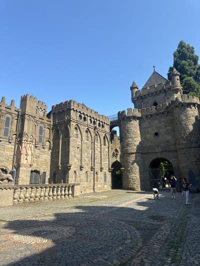 The image shows the Löwenburg castle in Kassel, Germany. The castle is made of stone and has a medieval appearance. In the foreground, there is a cobblestone courtyard with some people walking around. The sky is blue and clear. A person is bending down to pick something up from the ground. Another person is standing and looking at their phone. A third person is taking a photo. The architecture of the castle is elaborate, with turrets, arches, and decorative stonework. There is a large tree behind the castle to the right.