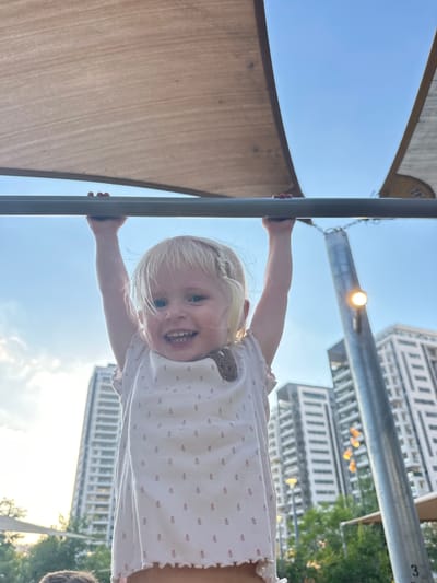 A young Caucasian girl with blonde hair and blue eyes is hanging upside down from a metal bar outdoors. She is smiling widely, showing her teeth, and appears joyful. She is wearing a white sleeveless top with a delicate pink floral pattern and ruffled edges. Her arms are bent as she grips the bar.

The background shows a bright blue sky with some clouds and the sun shining. In the distance, there are modern white buildings with many windows, indicative of an urban setting. There are also trees visible, suggesting a park or a green area within a city. A large beige sunshade or sail is visible in the upper left corner of the image, and another metal pole runs horizontally in the mid-ground, from which a light fixture is suspended.

The low angle perspective of the shot emphasizes the girl's inverted position and her cheerful expression. The overall mood is playful and lighthearted. The image was taken in Givat Shmuel, Israel, and the time of day appears to be daytime, likely late morning or afternoon, given the bright sunlight. There is no visible text in the image.