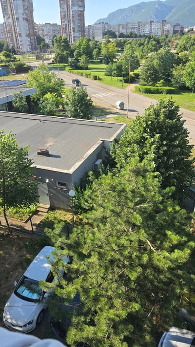 This media file is an aerial view of a residential area in Vratsa, Bulgaria. The scene depicts a street lined with tall, concrete apartment buildings on the left, and a lush green park with mature trees on the right. A road runs through the middle of the scene, with a dark-colored car visible driving on it. In the foreground, a low-rise building with a dark, flat roof is visible, with solar panels on its surface. A tall pine tree with dense green needles dominates the right side of the frame, casting dappled shadows on a white car parked below it. The time of day appears to be daytime, with bright sunlight and clear blue skies, indicating fair weather. The overall impression is one of a typical urban landscape in a European city, with a mix of residential buildings and natural elements. No people are clearly visible in the image.