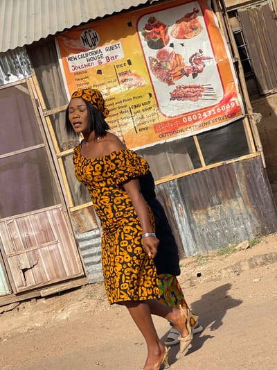 This image captures an outdoor scene in Unguwar Gabas, Nigeria, under direct sunlight. A woman with medium complexion is depicted mid-stride, facing slightly right with her mouth slightly open. She wears an off-the-shoulder, knee-length dress and a matching headscarf, both featuring a black background with irregular orange and yellow patterns. Her short, dark hair is visible beneath the scarf. She wears light-colored, pointed-toe high heels and a silver-toned bracelet on her left wrist.
Behind her, partially obscured by shadows and her body, a second individual is visible, wearing a black upper garment, a multi-colored patterned skirt, and light-colored sandals. Both stand on a light-colored, unpaved ground surface.
In the background, a building constructed from weathered corrugated metal and wooden panels is visible. Attached to this structure is a large, slightly torn orange and white banner. The banner reads "NCGH NEW CALIFORNIA GUEST HOUSE Kitchen & Bar," listing various food items such as Rice, Beans, Sema, Abacha, and different types of soup. It also displays images of assorted Nigerian dishes, including grilled fish, skewers, and cooked meats, and provides contact information: "08024316049" and "OUTDOOR CATERING SERVICES." Strong shadows are cast towards the lower left, indicating the sun's position.