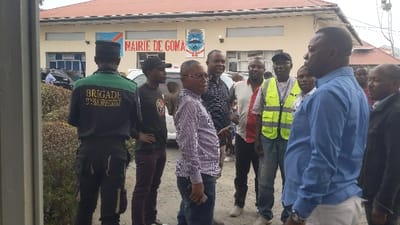 A group of men stand in front of the "Mairie De Goma" building, displaying a Congolese flag. One man is facing away, wearing a uniform with "BRIGADE ASSAINISSEMENT" on the back. Several men are in casual clothing, including button-down shirts and jeans. Some are wearing reflective vests. A few are turned to face the man in the blue shirt. The building behind them is light-colored with windows. A bush is present to the left side.
