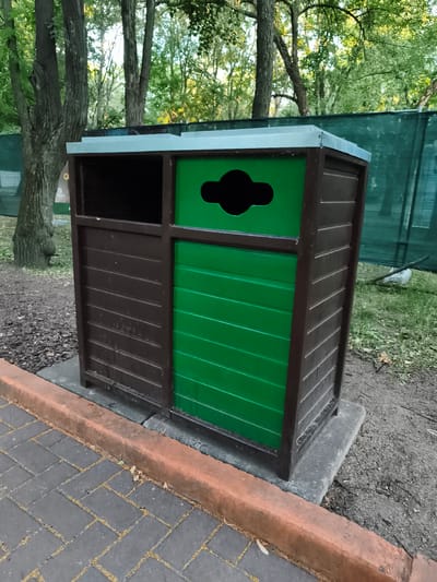 This is a medium shot, eye-level, outdoor photograph of a two-compartment trash receptacle. The receptacle is made of dark brown wood-like material with horizontal slats. The left compartment is open at the top and appears to be for general waste, with a dark interior. The right compartment is painted bright green and has a distinct, cutout opening at the top shaped like a stylized hourglass with a rounded middle section, commonly used for glass recycling. Both compartments have a light blue metal lid that overhangs slightly. The receptacle is situated on a concrete base.
In the foreground, a textured walkway made of dark grey rectangular pavers is visible, bordered by an orange-brown curb. Beyond the curb and to the right of the receptacle is a patch of grey dirt and gravel, with some dry leaves scattered on the ground. Further back, behind the receptacle, is a lush green lawn dotted with more dry leaves and some fallen branches. A tall, dark green mesh fence runs horizontally across the background.
Dominating the upper portion of the image are the trunks and canopy of several mature trees with green leaves, suggesting a park or wooded area. The lighting indicates it is daytime, possibly late afternoon, as the sunlight filters through the leaves, creating dappled shadows. The overall scene appears calm and natural, suggesting a public outdoor space that is maintained for waste disposal and recycling. There is no visible text in the image.