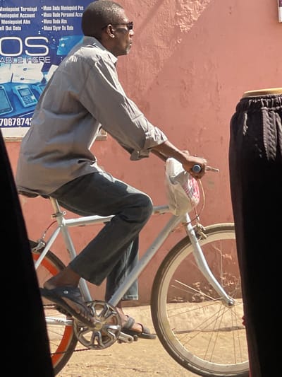 An adult male, dressed in a light grey long-sleeved shirt and dark grey trousers, rides a light-colored bicycle in Samaru, Nigeria. He wears dark sunglasses and dark sandals. A white plastic bag is suspended from the bicycle's handlebars, which have light blue grips. The bicycle features a white front wheel rim and an orange-brown rear wheel rim. The man's feet are positioned on the pedals, which include a silver-colored sprocket. The background features a textured, light reddish-pink wall. To the left, a partially visible blue sign advertises "Moniepoint Terminal," "Moniepoint Account," and "Sell Out Data," along with phrases in Hausa (e.g., "Muna Bada Moniepoint Pos") and images of point-of-sale devices. A partial phone number, "7067874," is also visible on the sign. A blurry, dark brown object with a ribbed texture is present in the extreme right foreground. The scene is illuminated by strong sunlight, casting shadows on the unpaved ground.