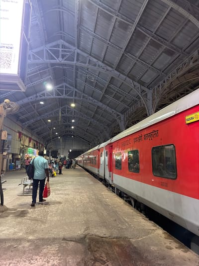 This image depicts a train at a station platform. The train cars are red and white, with "MADURAI" written on a yellow sign on the side of one car, indicating the destination. The platform is made of concrete and appears wet in places. A man in a light blue shirt and dark pants is walking away from the camera, carrying a black bag over his shoulder and a red shopping bag in his hand. Other people are visible on the platform, some sitting on benches, others standing. The station has a large, arched, metal roof supported by intricate trusses. Overhead lights illuminate the scene. In the background, there are shops and stalls. The architecture suggests an older, established train station. This is likely Chennai Central Railway Station in Chennai, India, a major transportation hub in the city.