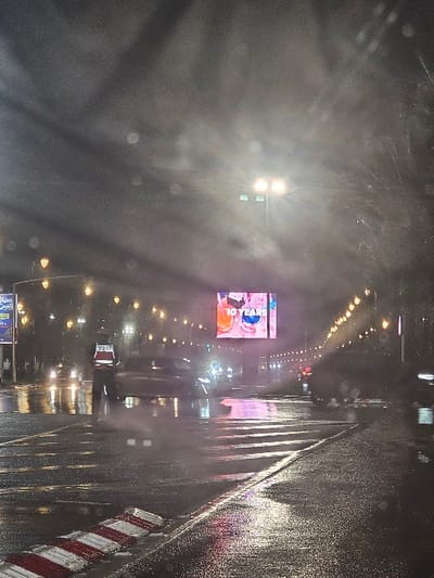 The image displays a nighttime street scene in Salé, Morocco, characterized by a wet road surface indicative of recent or ongoing rain. The central focus is a multi-lane asphalt road, highly reflective of ambient light sources.

In the immediate foreground, a red and white striped traffic calming device or barrier is positioned along the right edge of the visible road. A human figure, possibly traffic personnel, stands on the left side of the road, wearing a white uniform with red markings on the upper garment and dark lower attire. Multiple vehicles are present on the road, exhibiting motion blur, particularly a dark sedan in the mid-ground, indicating active traffic flow. Headlights and taillights from these vehicles illuminate parts of the wet pavement.

Overhead and lining both sides of the street are numerous streetlights, emitting a warm yellow-orange light and creating elongated reflections on the wet ground. A large, brightly illuminated digital billboard dominates the mid-ground, displaying an advertisement featuring colorful beverages and the English text "10 YEARS." To the left of this digital billboard, another static billboard with visible Arabic script is present. The background shows a continuation of the street, receding into the distance with further streetlights and indistinct building outlines. The sky is dark and appears overcast, with visible light flares originating from the streetlights.