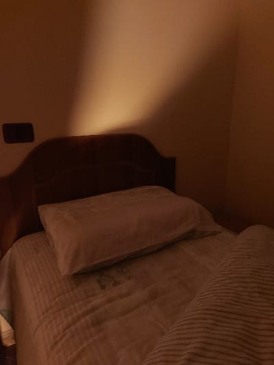 The image displays a dimly lit bedroom scene, located in Tiranë, Albania, likely captured during the evening or night as suggested by the warm, artificial lighting. A bed occupies the right side of the frame, featuring a light-toned duvet with subtle vertical stripes and a plain, light-colored pillow. A dark, possibly discolored or textured, patch is visible on the duvet beneath the pillow. Behind the bedding, a dark wooden headboard is partially visible, extending along the right edge. The left side of the scene is dominated by a plain, light-colored wall, which is bathed in a warm, golden glow emanating from an out-of-frame light source, presumably a bedside lamp. A small, dark rectangular object, resembling a wall outlet or a mobile phone, is mounted low on this illuminated wall. The scene is static, with no individuals or activities present. No text is visible within the frame.