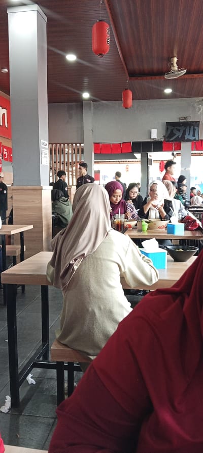 The image shows the interior of a restaurant in Katapang, Indonesia (latitude: -7.026, longitude: 107.549). The restaurant has a wooden ceiling with red lanterns hanging from it. There are four people sitting at a table eating. The people are all wearing clothing typical of Indonesia. The table is made of wood and has a variety of items on it, including food, drinks, napkins, and a blue container. There is a large, white column to the left of the table and a person to the right of the table out of frame, wearing a red shirt. The table is next to a window looking out onto a street. There is a white ceiling fan to the right. The scene is bright and well-lit. The restaurant appears clean and well-maintained. The atmosphere is relaxed and casual.
