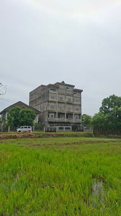 The image shows an unfinished, multi-story concrete building situated next to a lush green rice field. The building, which appears to be several stories high, has numerous windows and balconies, but its construction is incomplete, with exposed concrete and unpainted surfaces. A white van with red and white lettering, possibly an ambulance, is parked in front of the building. Dense green trees are visible behind and to the side of the building, adding to the natural setting. The rice field stretches to the right of the frame, with some water visible within it, indicating it is actively being cultivated. The sky is overcast, suggesting a cloudy or perhaps a humid day. The image is oriented vertically, with the sky at the top and the ground at the bottom. The overall scene depicts a rural or semi-rural area in Kutaglumpang, Indonesia, with a juxtaposition of a large, unfinished structure against the natural landscape. There is no discernible activity or event taking place.