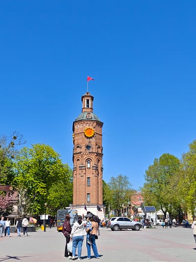 The image captures a sunny day in the city of Vinnytsia, Ukraine, focused on the iconic Water Tower. The brick tower, topped with a green dome and a red flag, stands prominently in the city center. A large clock is visible on the tower's façade.
In the foreground, several people are gathered on the paved square. One group is taking photographs of the Water Tower, with one person holding a small child. Other pedestrians are walking through the square in various directions.
Behind the group, a grey bus with Cyrillic lettering is visible. A silver SUV is parked on the square. Trees with lush green foliage frame the scene. In the background, other buildings and street furniture are present, including a small structure with a solar panel. The sky is a clear, bright blue.