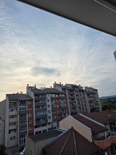 The image depicts a view of apartment buildings and rooftops in Gabrovo, Bulgaria. The scene is dominated by several multi-story residential buildings with light-colored facades and dark tiled roofs. The buildings are arranged in a row, with some showing balconies and windows. In the foreground, there are lower-lying buildings with red tiled roofs. The sky above is partially cloudy with visible contrails from aircraft. The lighting suggests it is either early morning or late afternoon. There are no people clearly visible in the image. The overall impression is an urban residential environment.