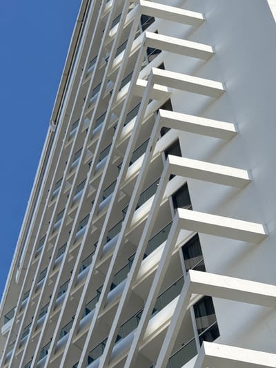 This media file is a low-angle, wide shot of a white, modern architectural structure with a repetitive, angular design. The structure appears to be a roof or canopy supported by numerous white, rectangular beams that angle upwards from a lower plane. Between these beams are horizontal sections that create a series of rectangular openings. Within these openings, dark-tinted windows are visible, suggesting an interior space beneath.

The structure fills the majority of the frame, extending from the top left to the bottom right. The repeating pattern of beams and openings creates a strong sense of depth and rhythm. Below the architectural element, a clear, bright blue sky occupies the lower portion of the image, indicating it is daytime and the weather is fair. The lighting suggests it is likely midday, with strong shadows cast by the architectural features.

No people are visible in the frame. The focus is solely on the geometric design and materials of the building. The overall impression is one of clean lines, modern design, and a sense of scale. The context of Santa Marta, Colombia, suggests a tropical or subtropical climate, which aligns with the clear blue sky. There is no visible text in the image.