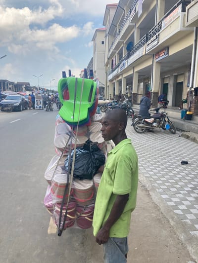 This image captures a bustling street scene in Umuode, Nigeria, during the daytime under an overcast sky. In the foreground, a dark-skinned man, appearing middle-aged, wears a light green long-sleeved shirt and blue jeans. He is focused on the goods he is transporting, looking downwards.
The man is pushing or pulling a cart heavily laden with various items. Prominently stacked on the cart are several bright green plastic chairs, along with others in red and purple, all wrapped in clear plastic. Below the chairs, numerous circular, flat items are visible, individually wrapped in plastic. These items appear to be mats or possibly hats, coming in shades of beige, maroon, and purple. A black plastic bag is also secured to the cart.
The background reveals a lively urban environment. To the left, a street stretches into the distance, populated by motorcycles and cars, indicating active traffic. Alongside the street are multi-story buildings housing various shops. Shop signs are visible, with readable text including "FASHION SHOP," "SISTERS," and "BEST OF FASHION." The sidewalk in front of these shops features a distinctive checkered pattern of light and dark tiles. Other people are visible further down the street, some on motorcycles, contributing to the busy atmosphere of a commercial district. The overall scene suggests a typical day of commerce and movement.