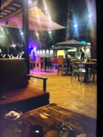 The image depicts an outdoor seating area of a restaurant or bar at night. The scene is illuminated by string lights and overhead lamps, casting a warm glow on the surroundings. In the foreground, a wooden table is visible with some objects on it, including what appears to be a candle in a holder. To the left, a plush chair with a dark cushion and a wooden frame is positioned. The background reveals more seating arrangements, including tables and chairs, as well as umbrellas for shade. There are also some figures blurred in the distance, suggesting patrons or staff. The overall ambiance is casual and inviting, hinting at a lively establishment. The location is identified as a city in Nigeria, specifically Udu.