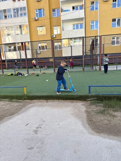 The image shows a boy riding a blue scooter on a green turf field. He is wearing a dark blue long-sleeved shirt and blue jeans. The field appears to be part of a playground or sports complex, with a fence bordering the turf and a building in the background. The building is a multi-story apartment complex with yellow and off-white sections, featuring many windows and air conditioning units.

Several other people are visible in the background, some standing and watching, others seemingly interacting. One person in a black and white patterned shirt is standing near the fence, while another person in a red and yellow soccer jersey is on the turf. There are also some sports equipment and bags scattered on the ground, including a black hat and a yellow backpack.

The scene suggests a casual outdoor activity, likely in the afternoon given the lighting. The weather appears to be clear. The presence of the apartment building and the artificial turf field are common in urban or semi-urban environments. The image is oriented vertically. There is no visible text in the image. The overall mood is one of everyday activity and outdoor recreation.